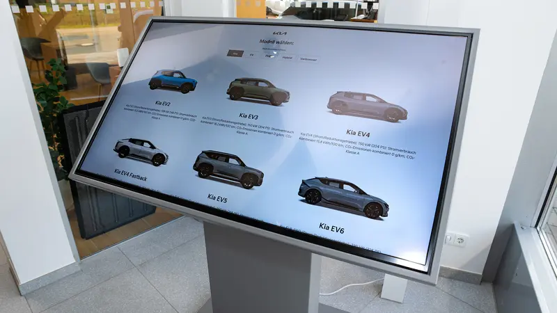 Interaktiver Bildschirm im Showroom zeigt sechs Kia Elektrofahrzeugmodelle mit kleinen Abbildungen und Modellnamen.