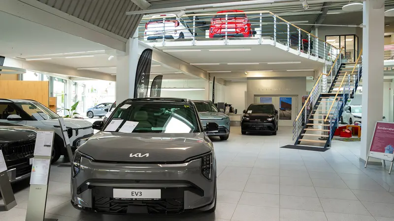 Innenansicht unseres Showrooms mit mehreren ausgestellten Kia und BYD Fahrzeugen, im Vordergrund ein Kia EV3, im Hintergrund eine Treppe zu einer Galerie mit weiteren Autos.