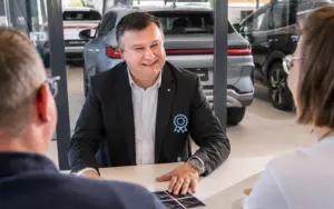 Verkäufer Valeri Dimmel sitzt im Anzug lächelnd an einem Tisch und spricht mit zwei Personen in einem hellen Verkaufsraum mit großen Fenstern