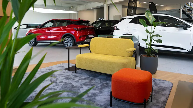 Showroom mit einem roten und einem weißen Kia Modell, gelber Couch, orangem Hocker und grünen Pflanzen auf grauem Teppich