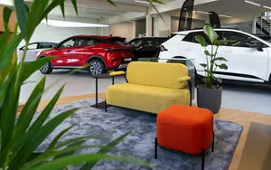 Showroom mit einem roten und einem weißen Kia Modell, gelber Couch, orangem Hocker und grünen Pflanzen auf grauem Teppich
