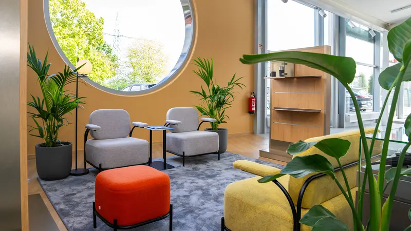 Moderne Lounge mit runden Fenster, grauen Sesseln, orangem Hocker, gelbem Sofa, Pflanzen und grauem Teppich in unserem Verkaufsraum-Wartebereich